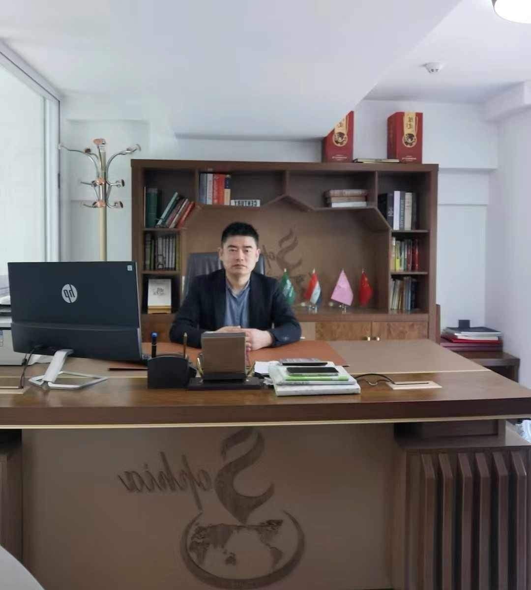 Man sitting at an office desk with bookshelves and a laptop, surrounded by flags and books.