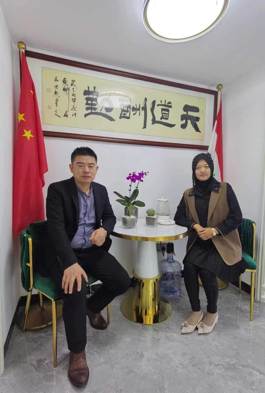 Two people seated at a small table with Chinese calligraphy and flags in the background.