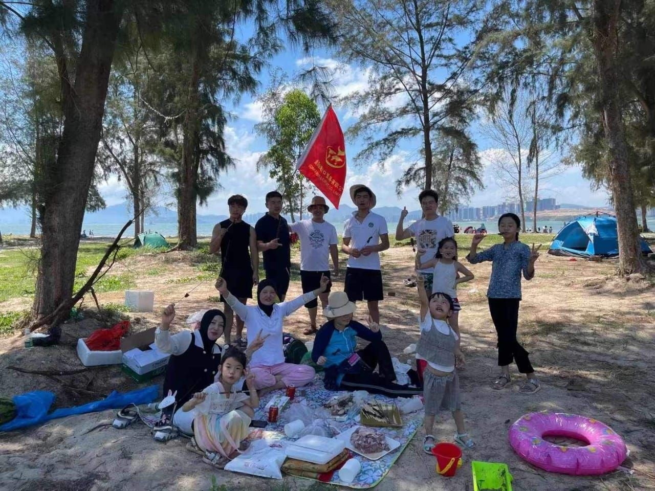 Group of people enjoying a picnic under trees, with tents and beach in the background.